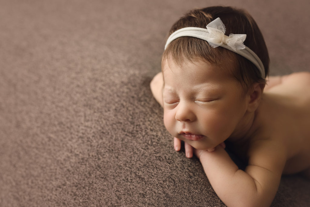 newborn baby girl with white bow on headband in face forward position on brown blanket