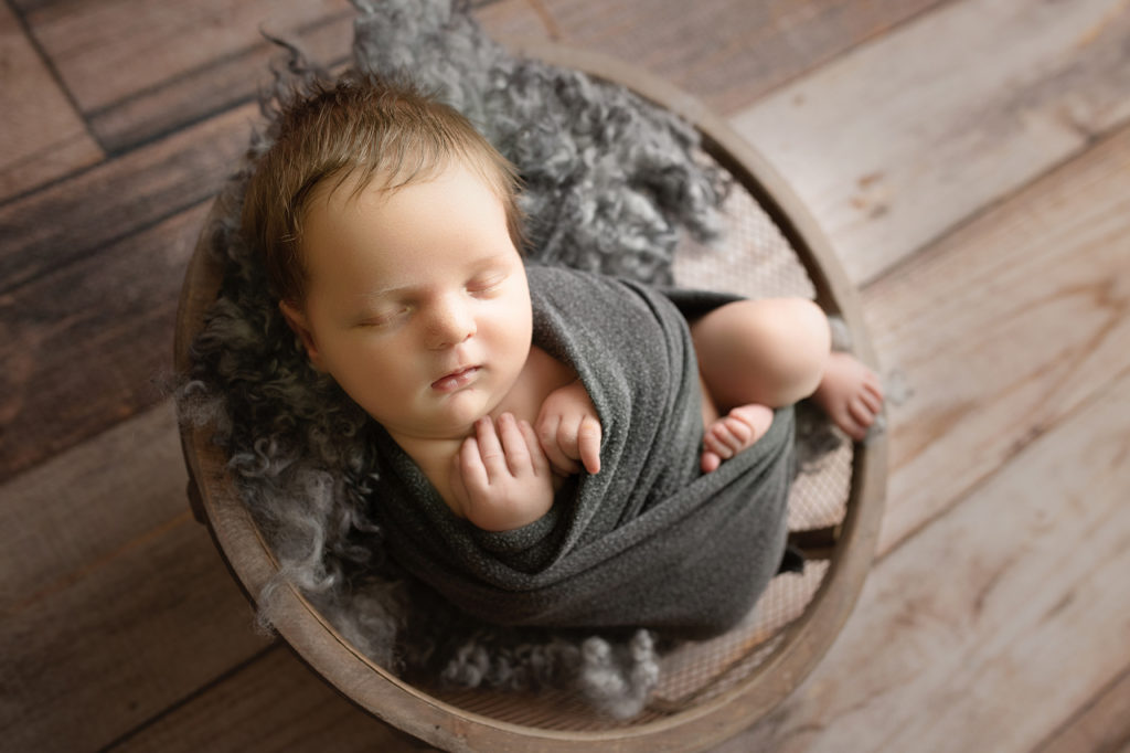 Newborn baby boy in brown mesh bowl and gray fur and wrap