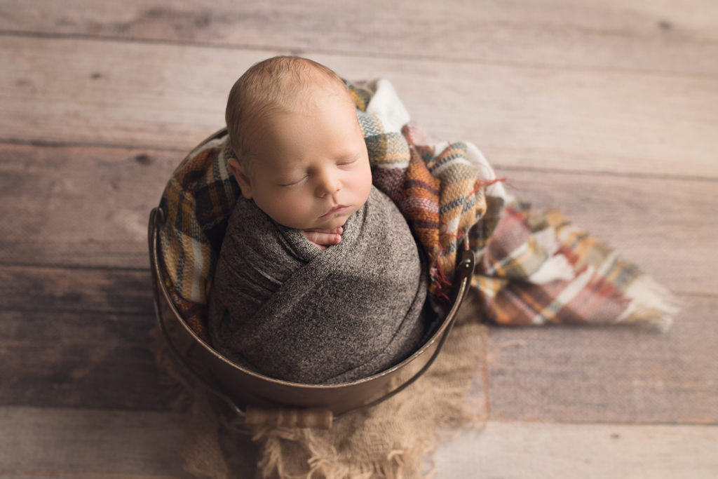 newborn baby boy in brown bucket wrapped in brown plaid