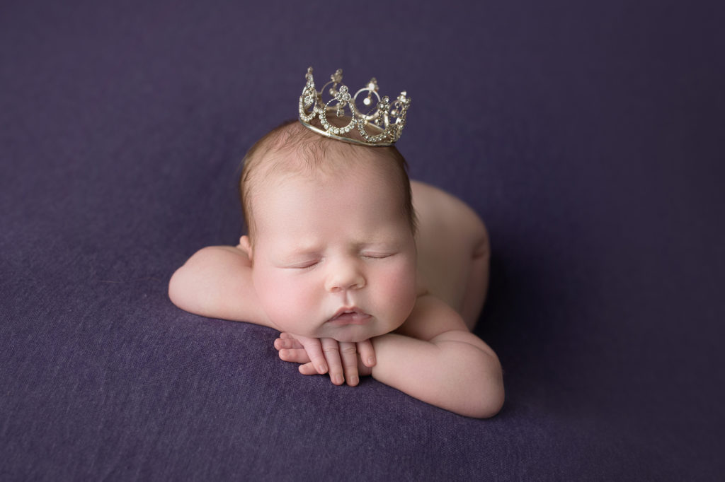 newborn baby girl face forward on purple blanket wearing a tiara