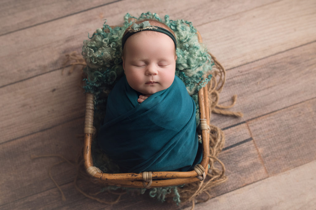 newborn baby girl in teal wrap with matching headband laying in a bamboo basket