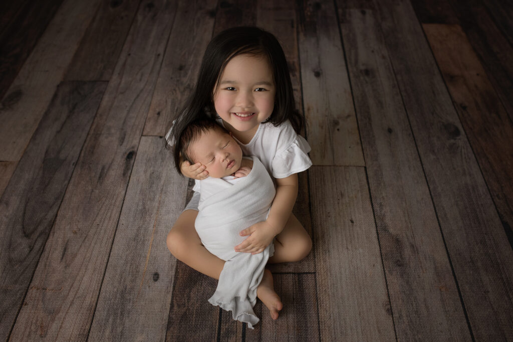 Newborn Baby and big sister in a sibling shot sitting up with a white wrap