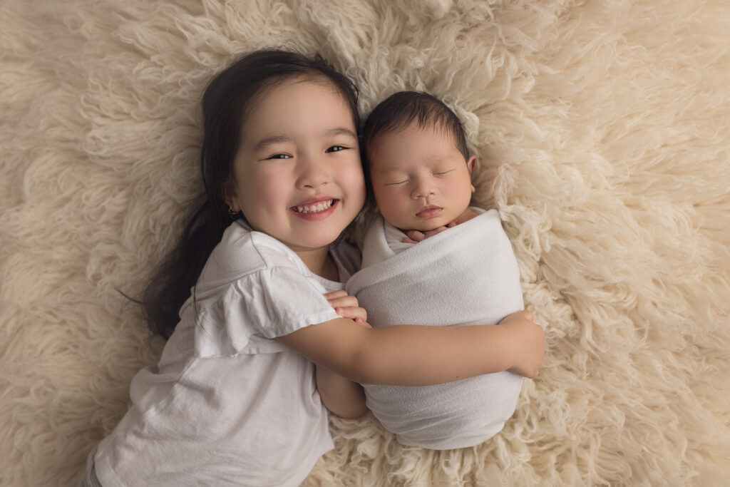 Newborn Baby boy being held tight by his big sister on a white blanket