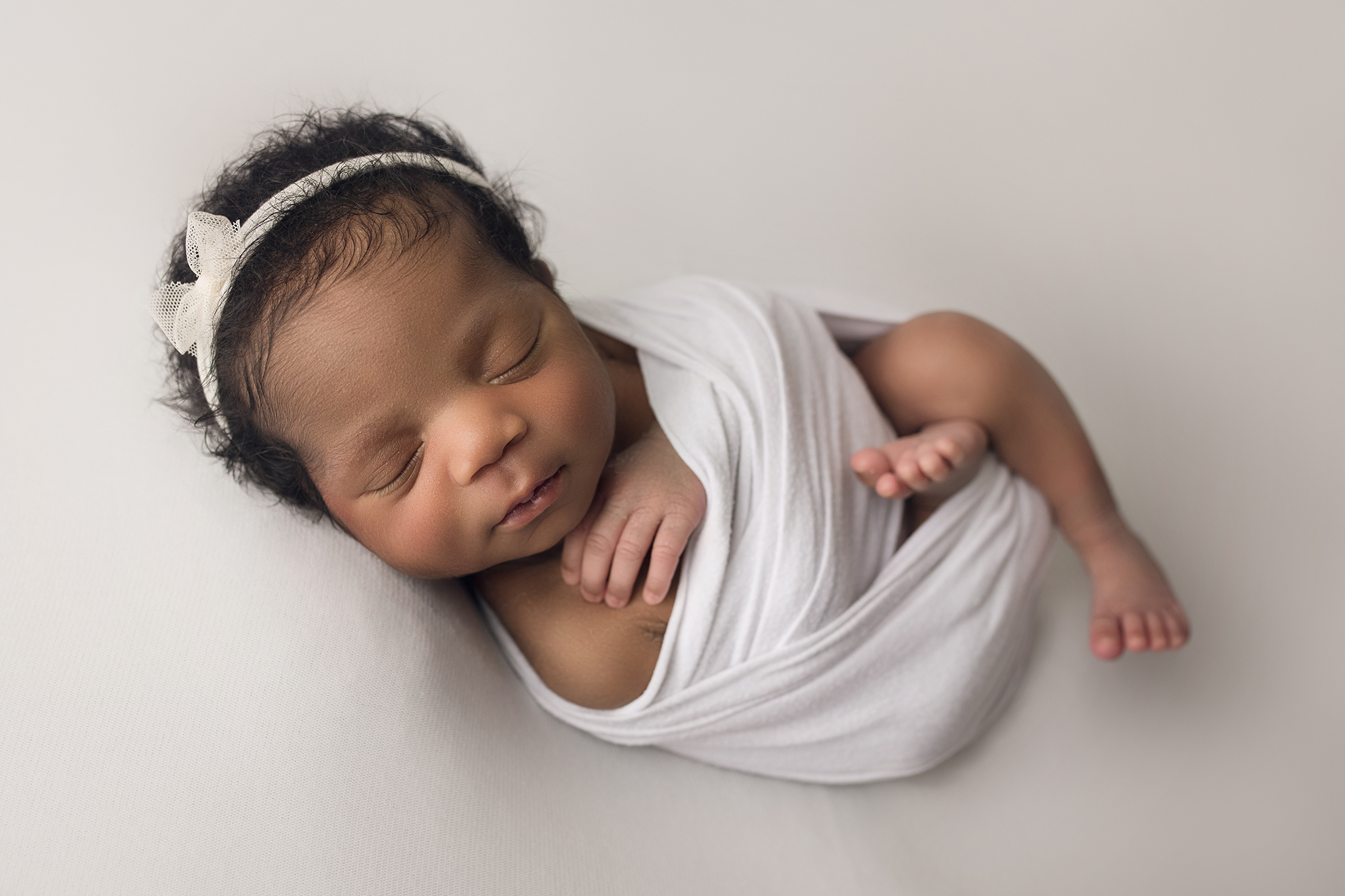 Newborn baby girl with white wrap and white bow laying on white blanket