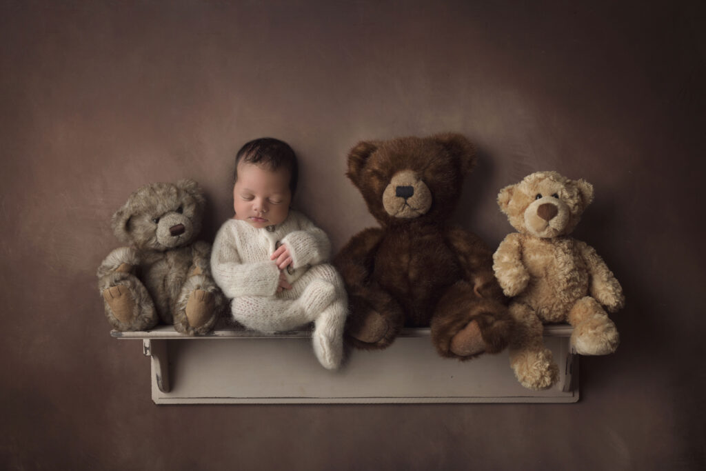 newborn baby boy sitting on a shelf with a bunch of teddy bears ina white knitted footed outfit