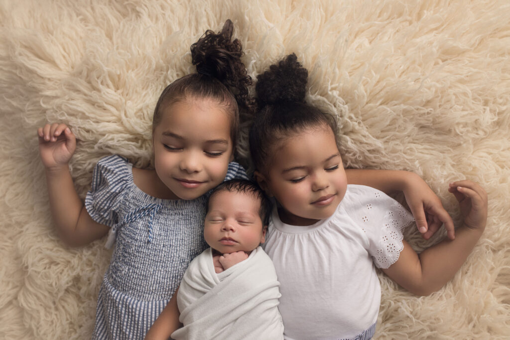 newborn baby boy wrapped in white being cuddled by his two sisters