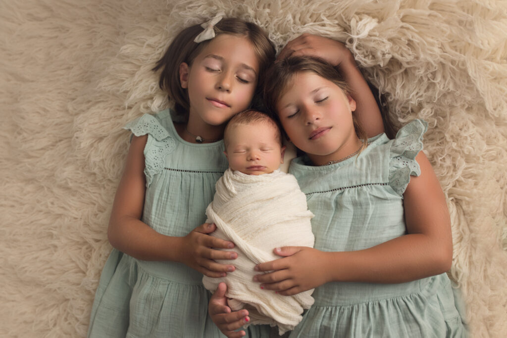 newborn baby boy laying on white fur laying asleep with two sisters