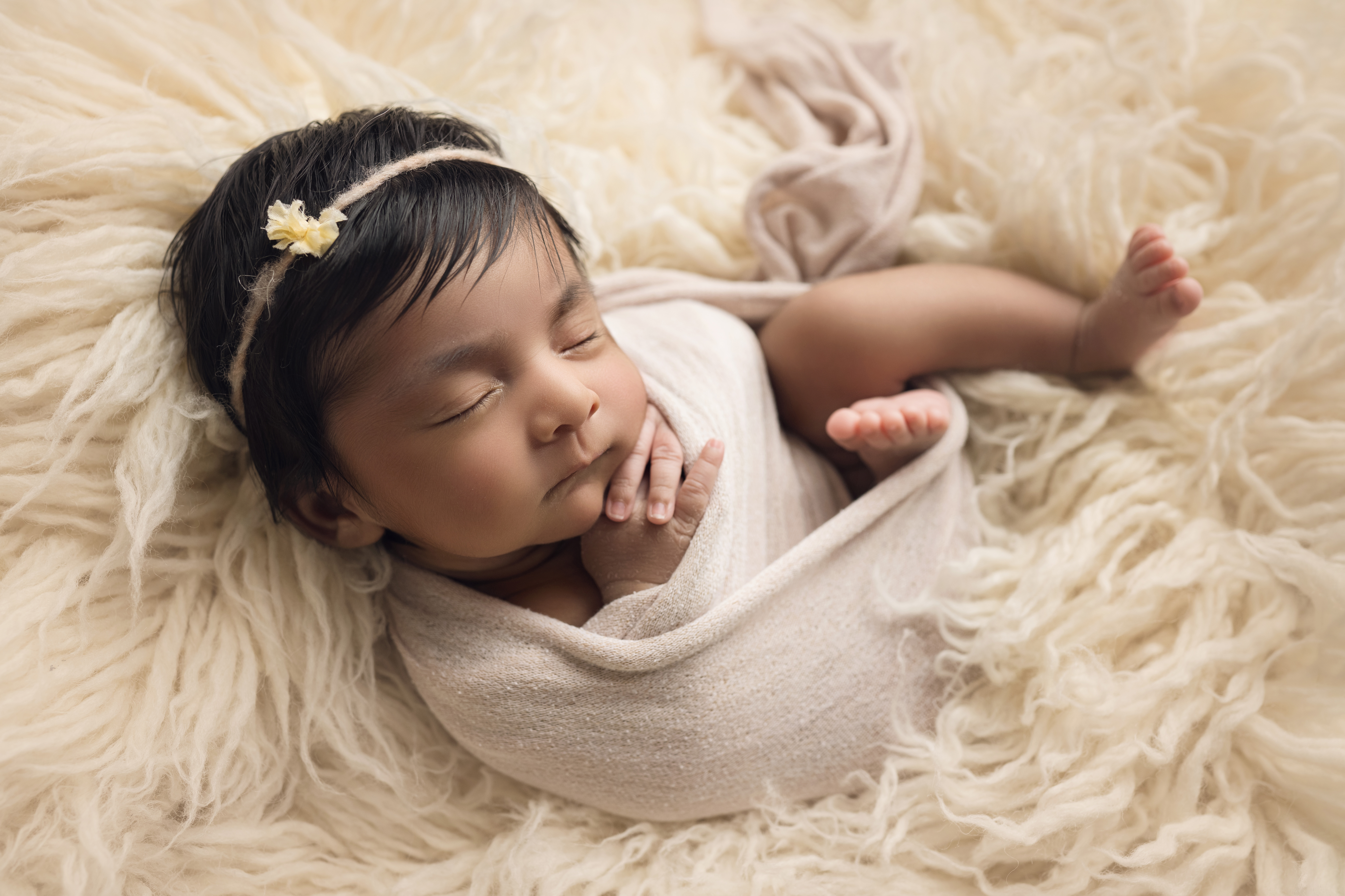 Newborn baby girl wrapped in cream with matching bow curled up on cream fur