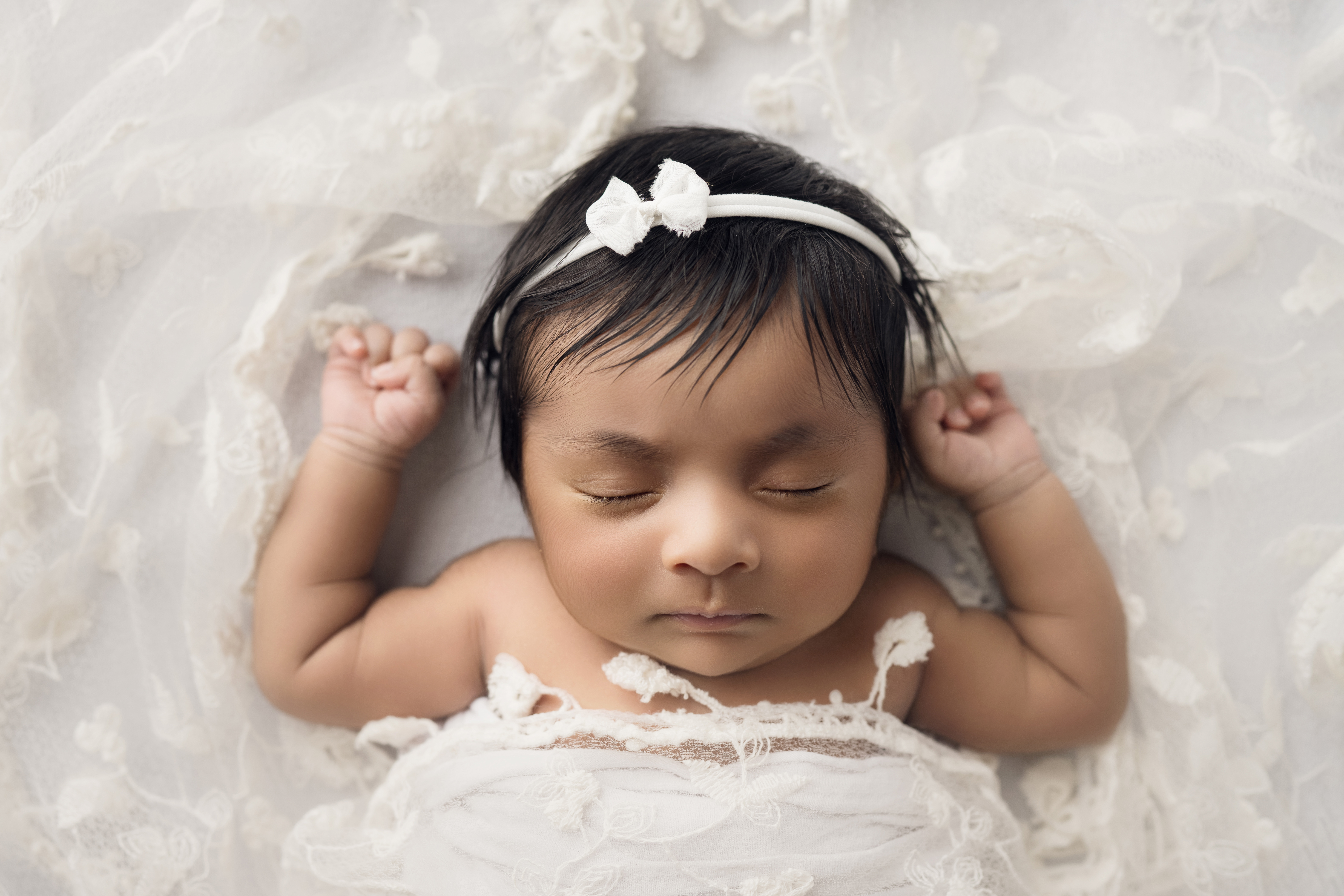 Newborn baby girl wrapped in white with matching bow on white blanket with lace