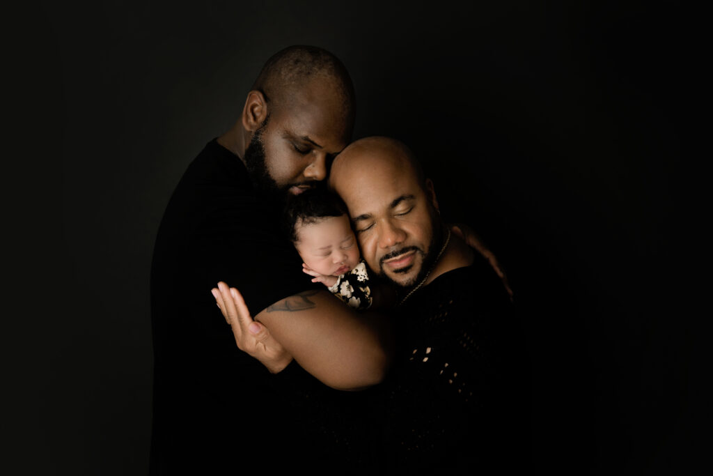 Newborn baby girl with her two dads snuggling on black backdrop