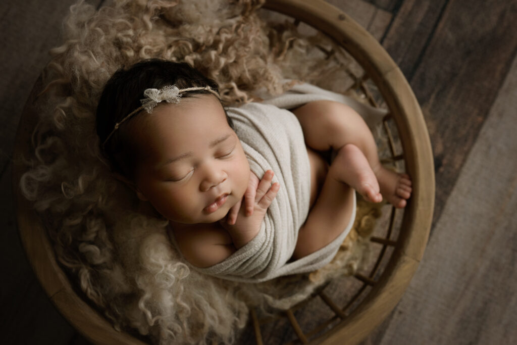 Newborn baby girl curled up in cream wrap in a bowl on a wood floor