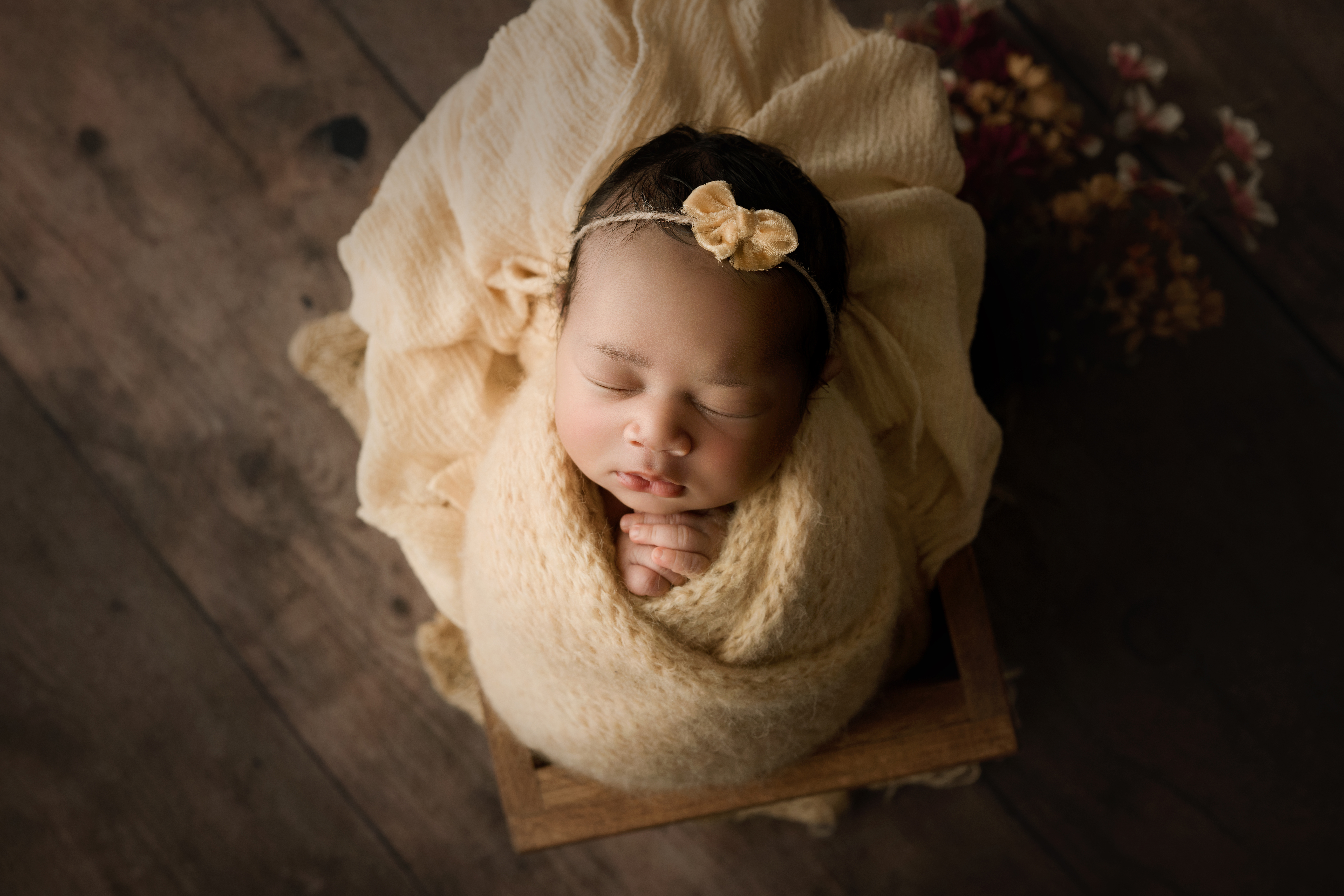 Newborn baby girl in yellow wrap with yellow headband laying in a box with flowers