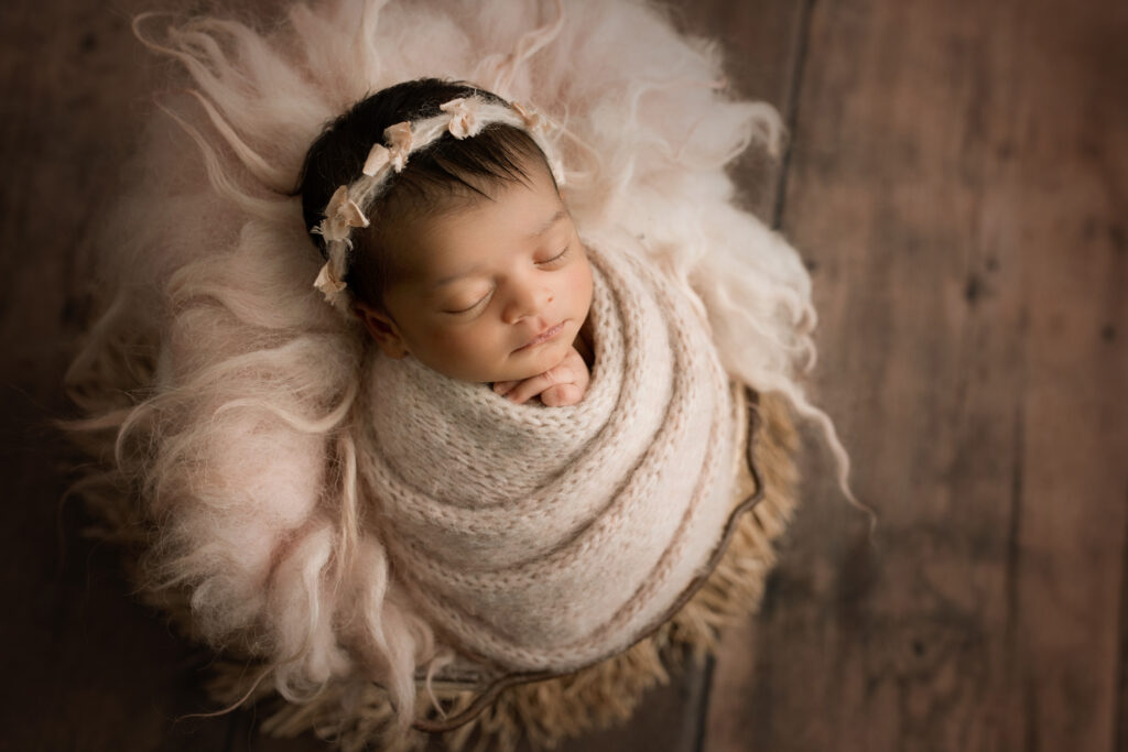 Newborn Baby Girl wrapped in a pink knit wrap with matching bow headband laying in a basket of fur