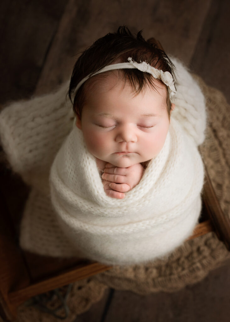 newborn baby girl wrapped in a cream knit blanket with a delicate headband, peacefully sleeping on a rustic wooden surface in a professional photography session. Holly Springs NC Newborn Photographer