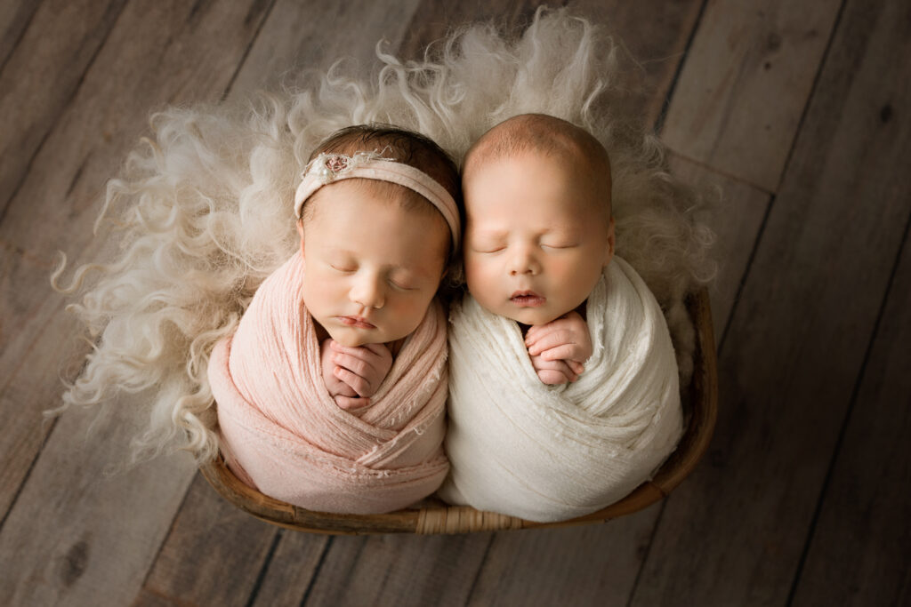 Newborn twins wrapped in pink and white blankets, sleeping peacefully in a rustic basket.