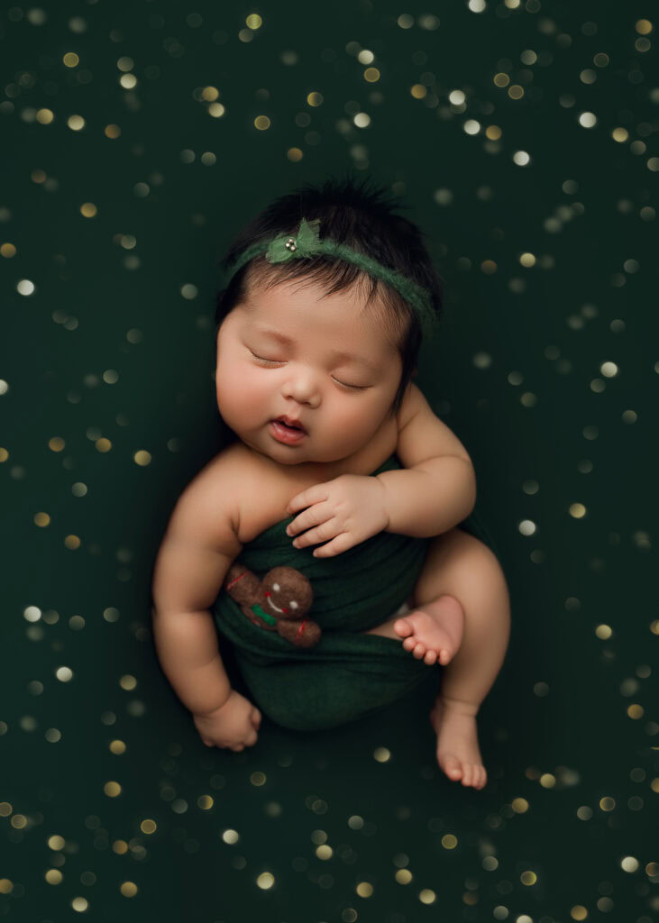 Newborn Baby girl wrapped in green Christmas Wrap on green blanket with green bow holding a gingerbreadman. Micro-Mini Christmas Newborn Session