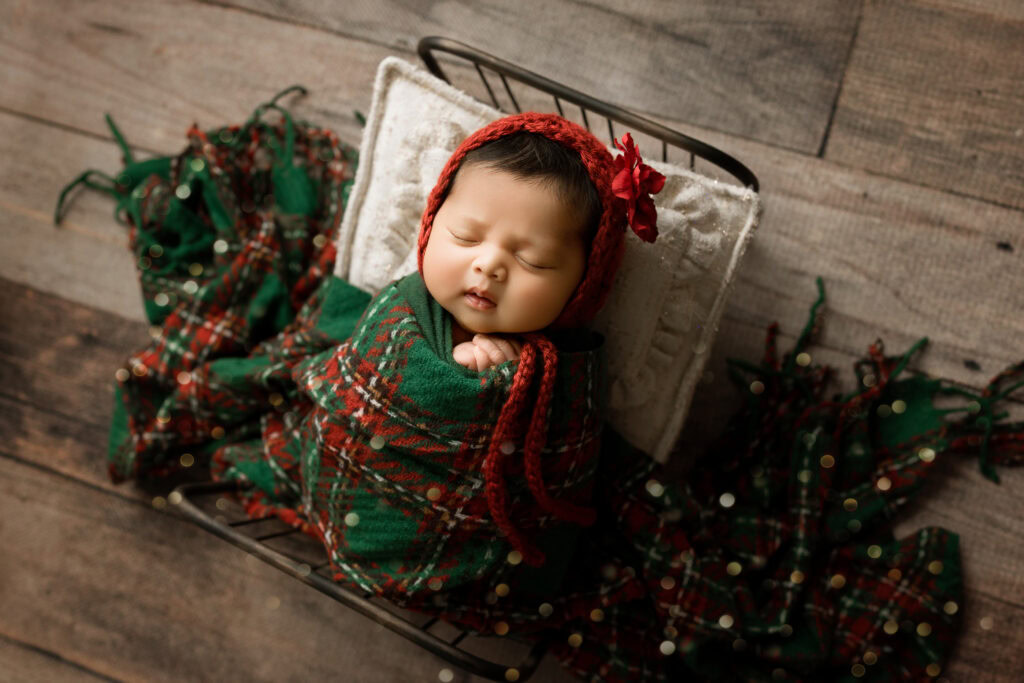 Newborn Baby girl wrapped in green Christmas Wrap in a bed with Christmas Plaid Blanket and christmas tree 