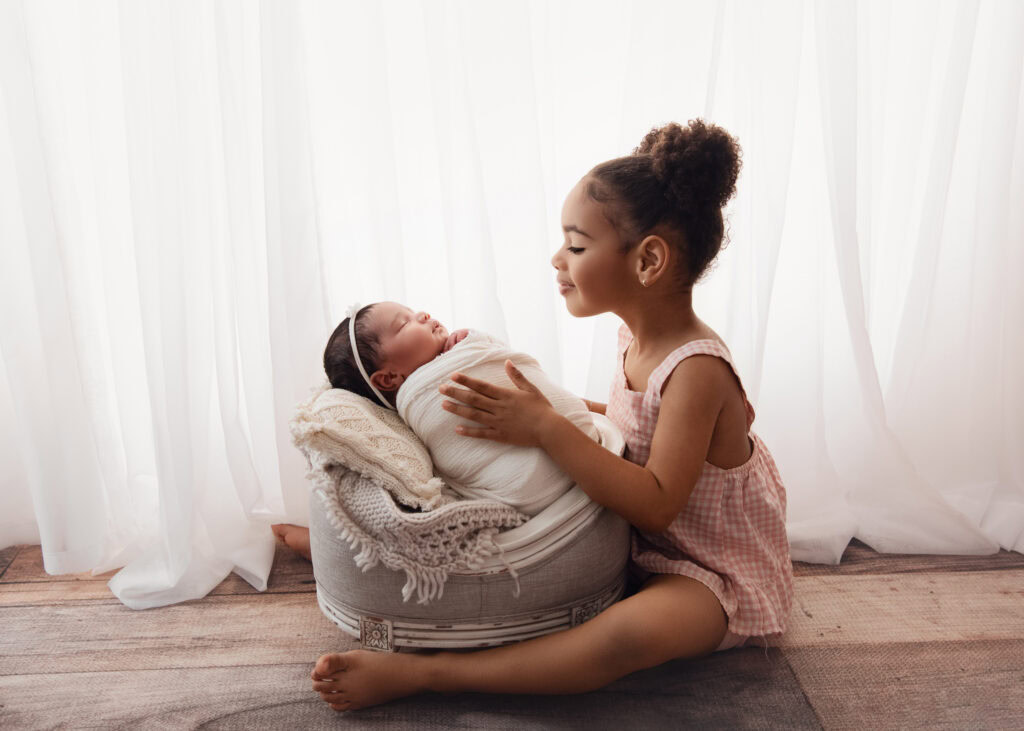 Newborn baby girl wrapped in a cream wrap and matching bow laying in a newborn crib being held by her big sister
