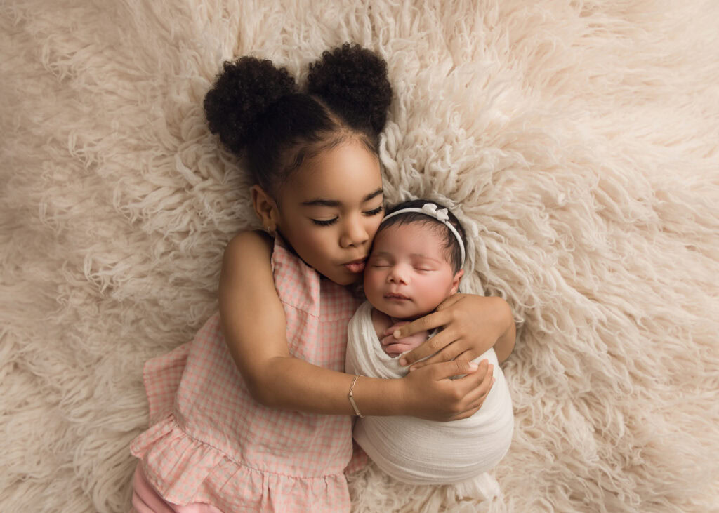 Newborn baby girl wrapped in a cream wrap and matching bowl being snuggled on a cream fur by her beautiful big sister