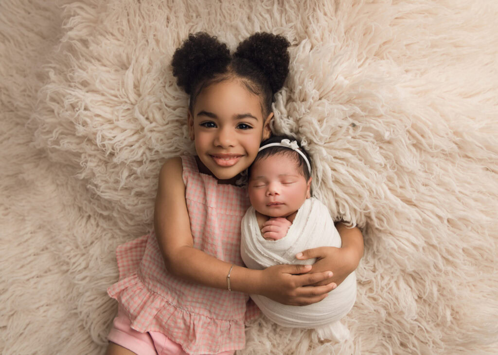 Raleigh Newborn photographer Newborn baby girl wrapped in a cream wrap and matching bowl being snuggled on a cream fur by her beautiful big sister