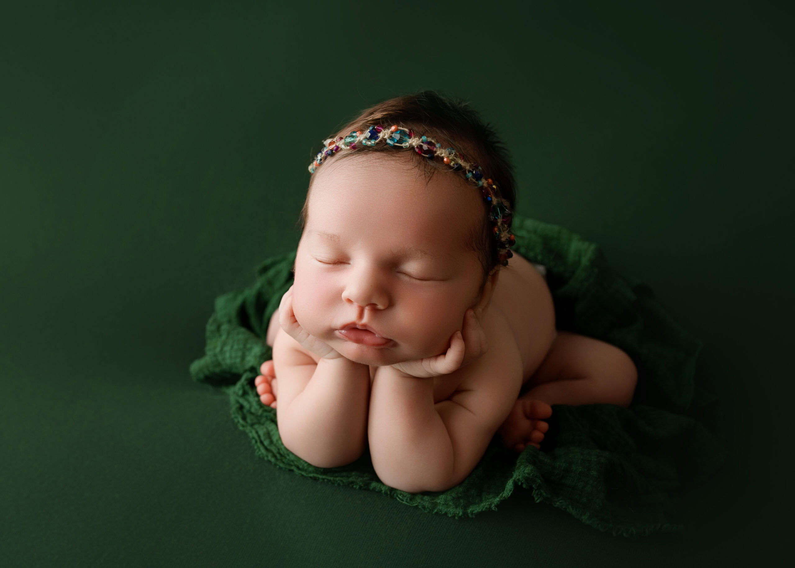 Newborn baby girl in the Froggy pose on a green backdrop with a headband made of multi colored gems